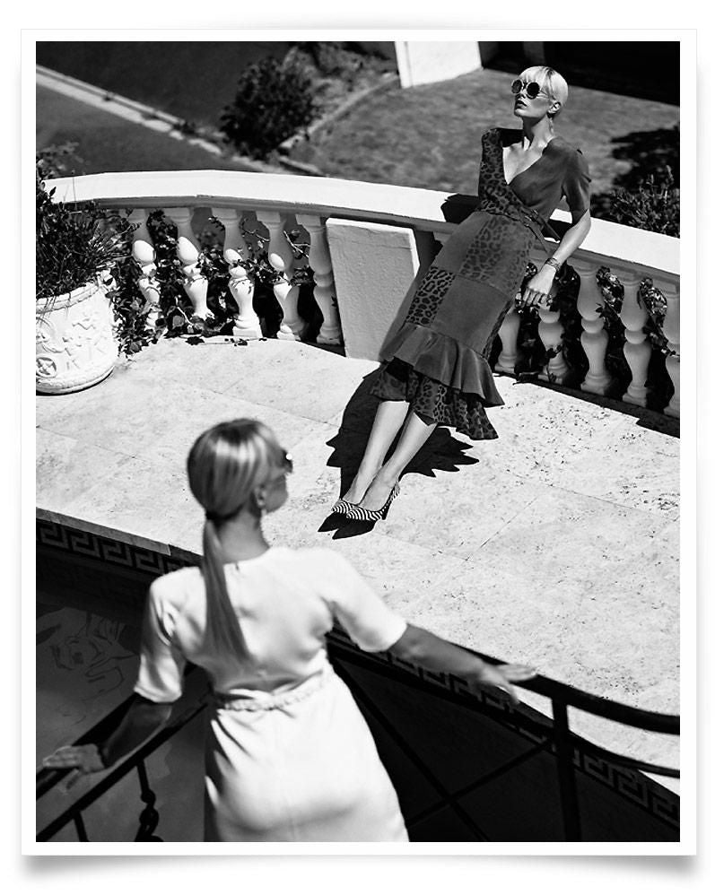 Two women with light hair and sunglasses are positioned on a sunlit terrace, one leaning against a balustrade, the other in the foreground.