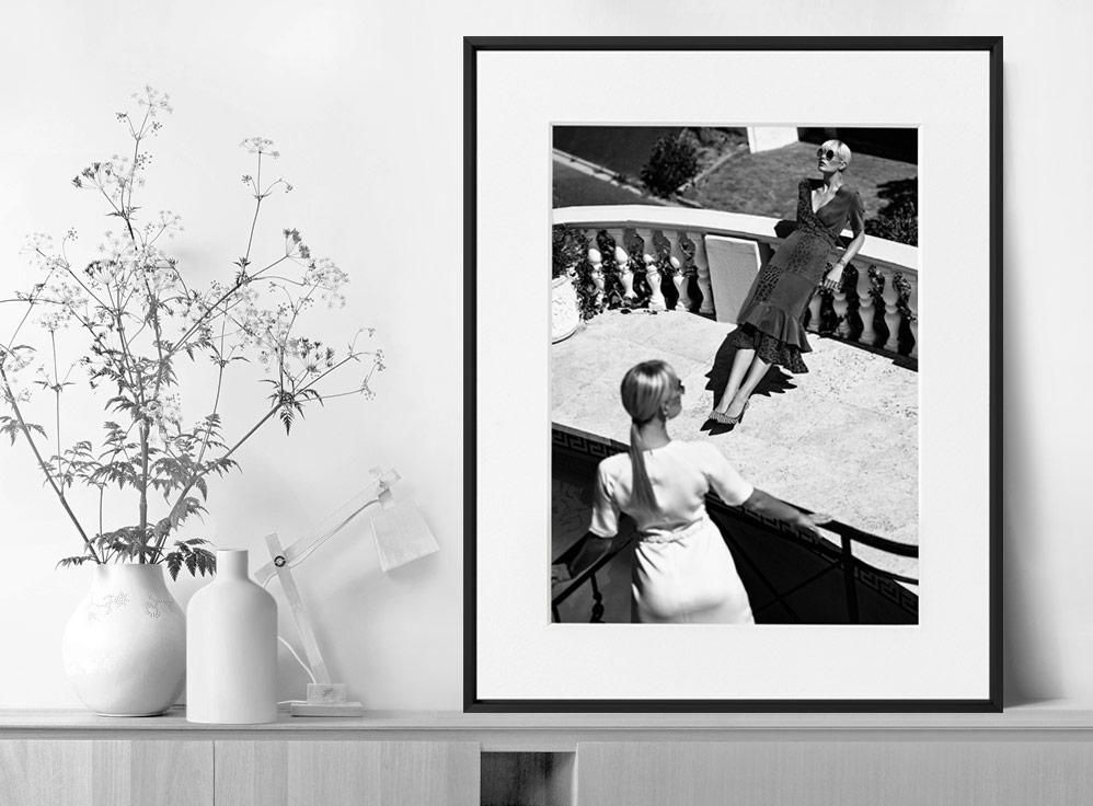 A framed black-and-white photograph shows two women on a sunlit terrace, one leaning on a balustrade and another standing below.