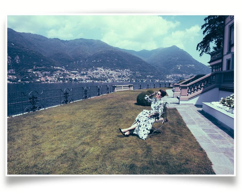 A woman in a patterned dress sits on a bench beside a lawn, with a lake and mountains in the background under a partly cloudy sky.