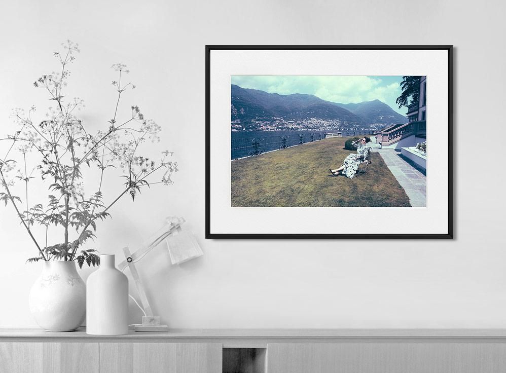 A framed color photograph shows a woman seated on a lawn chair beside a railing, with mountains and water in the background.