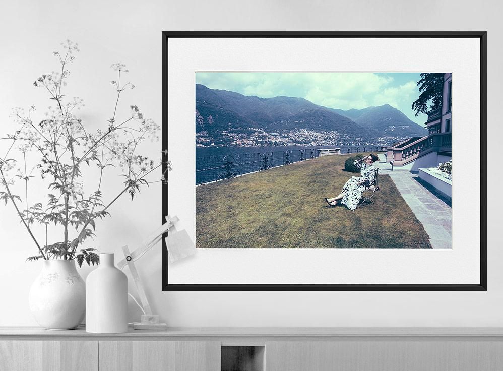 A framed color photograph shows a woman in a patterned dress seated on a lawn beside a lake, with mountains in the background.