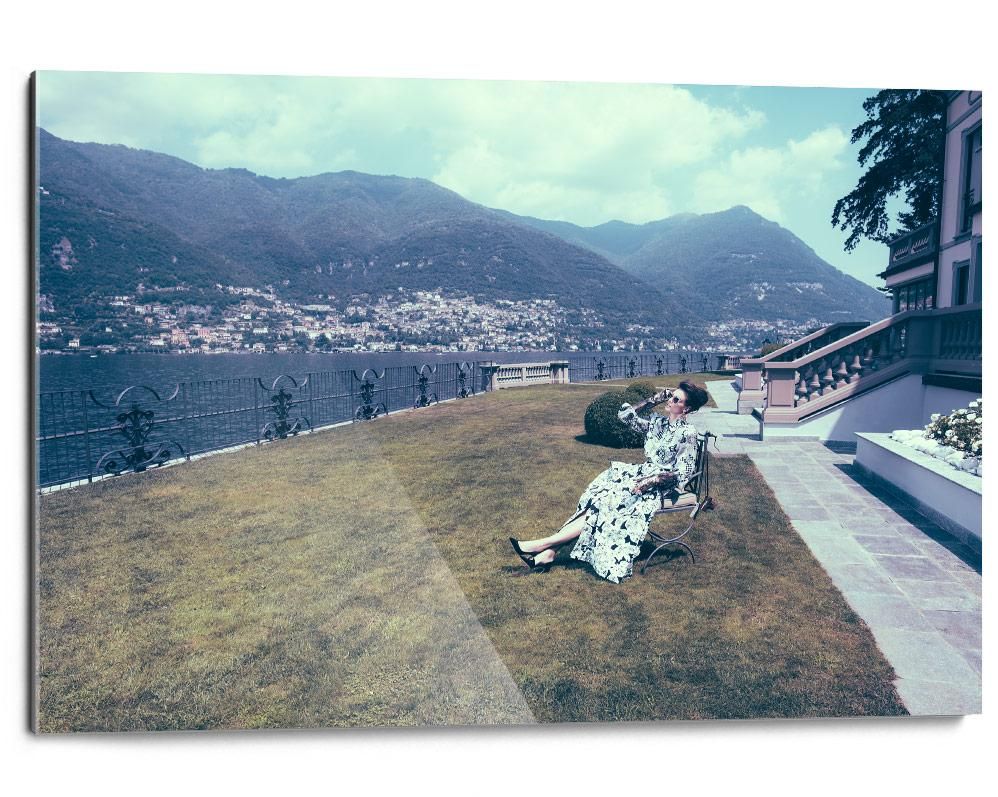 A woman in a patterned dress sits on a bench beside a large house, with mountains and a lake visible in the background.