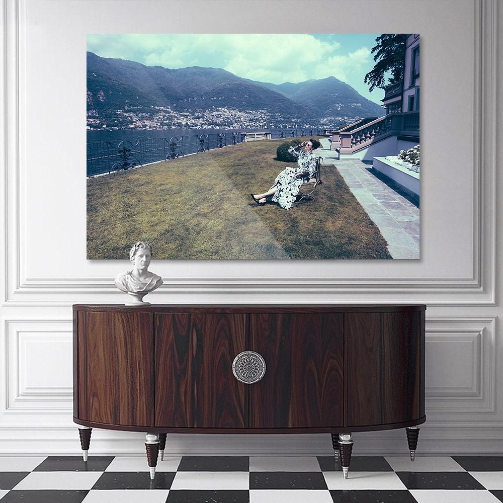 A framed photograph of a woman in a patterned dress seated on a lawn overlooks a lake and distant hills, mounted above a wooden cabinet.