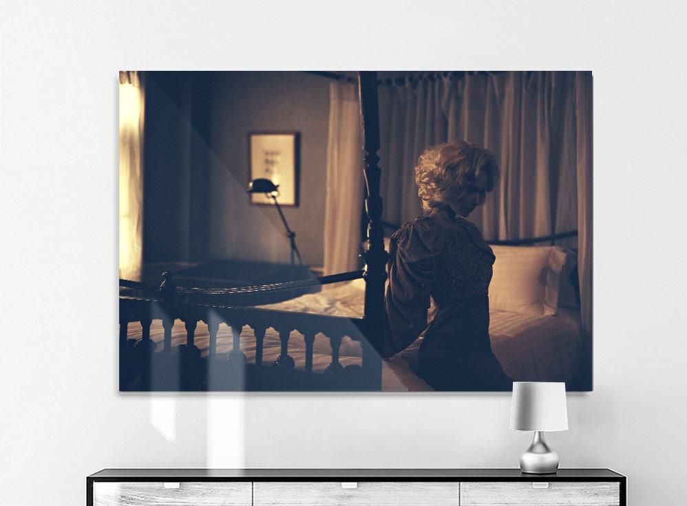 A woman with light hair sits on the edge of a four-poster bed in a dimly lit room with curtains and framed art on the wall.