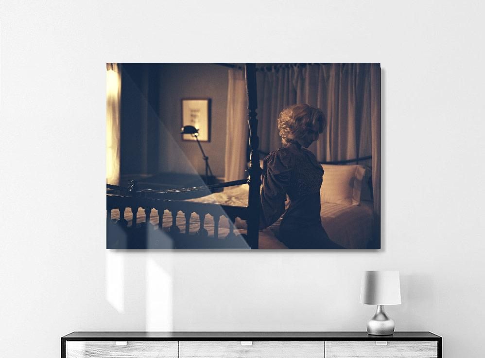 A framed photograph on a white wall shows a person with light hair seated on the edge of a bed in a dimly lit room.