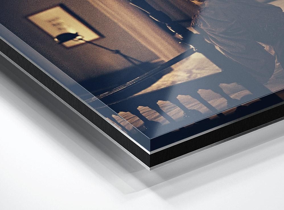 A cropped view shows a glossy print with a partial figure, a railing, and a blurred framed object on a wall in warm indoor light.