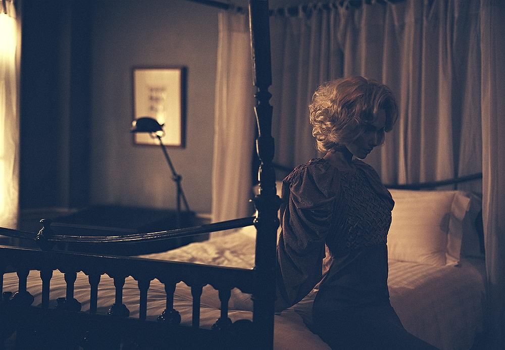 A person with light, wavy hair sits on the edge of a bed in a dimly lit room with curtains and a floor lamp in the background.