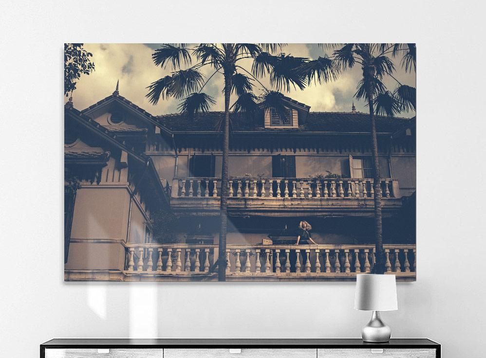Two tall palm trees partially obscure a two-story house with a balcony and balustrade under a cloudy sky. A person leans on the upper railing.