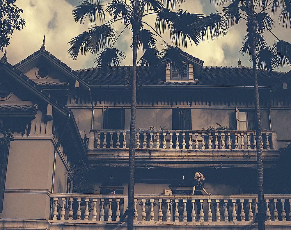 Tall palm trees partially obscure a two-story building with balconies and balustrades; a person is visible on the lower balcony.