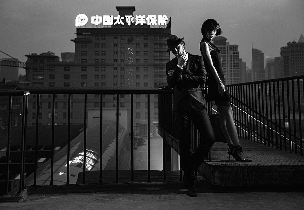 Two people in dark clothing pose on a rooftop walkway at dusk, with city buildings and a lit sign in the background.